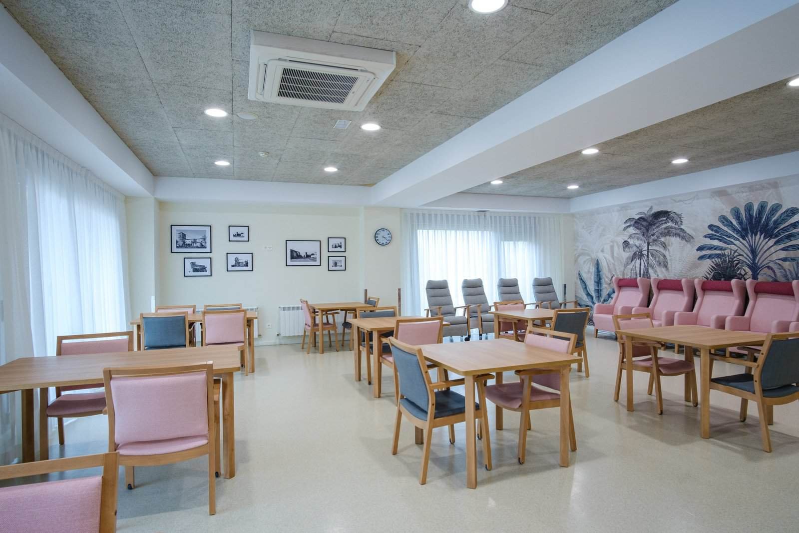 Sala común luminosa en la residencia Colisée Miñano, con mesas y sillas en tonos suaves, decoración mural y zona de descanso para residentes.