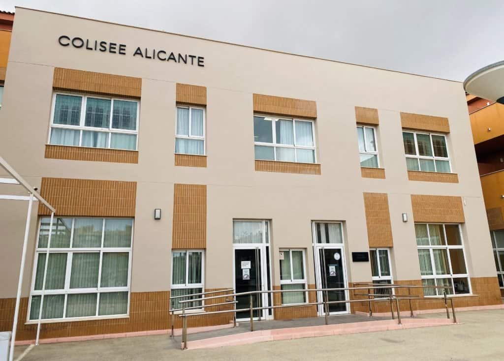 La fachada exterior del edificio de dos plantas de Colisée Alicante. Es una construcción moderna de tonos claros con un letrero en la parte superior que dice "COLISEE ALICANTE" y una rampa de acceso a la entrada principal.