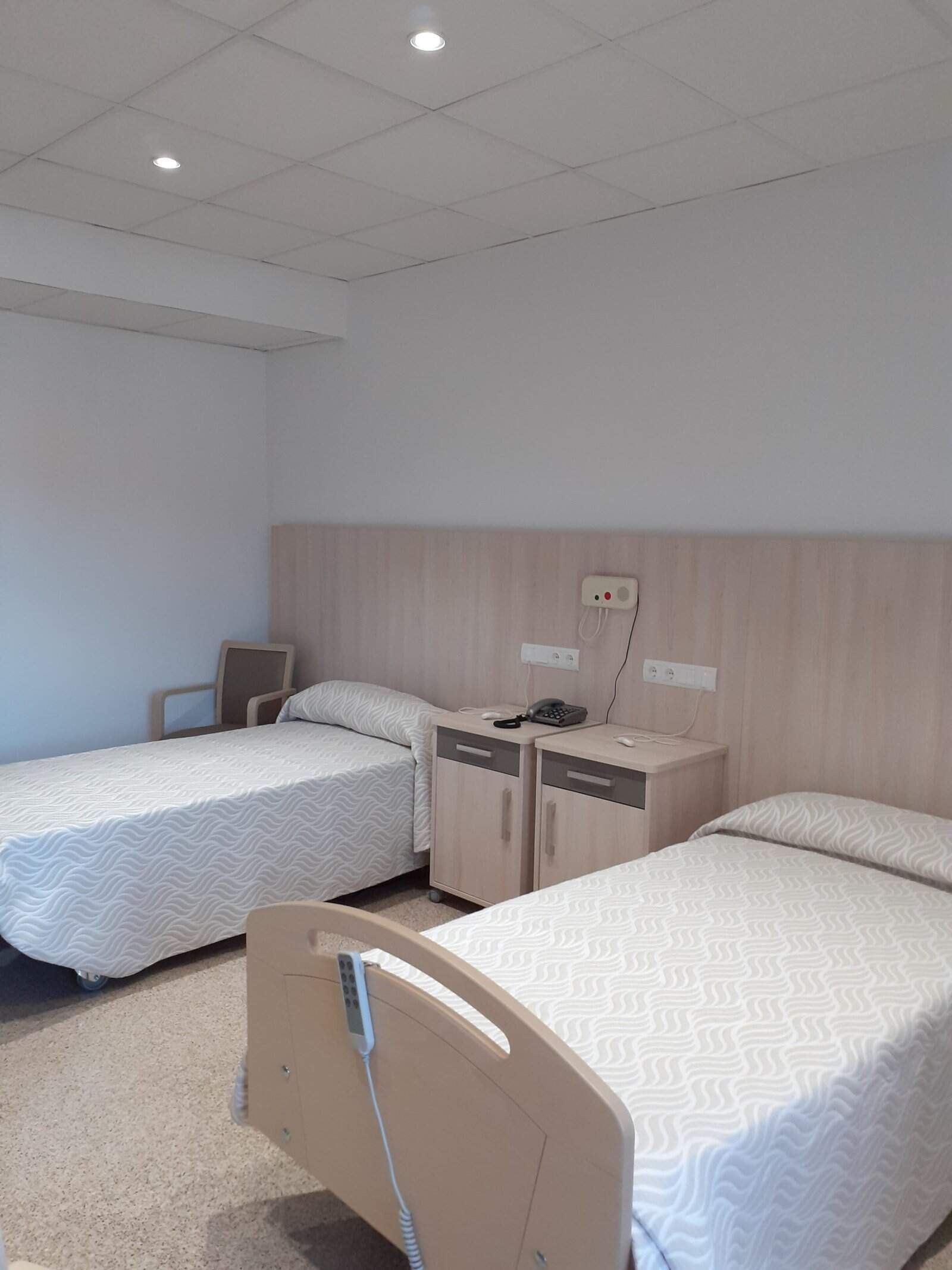 Un dormitorio doble, limpio y luminoso, en la residencia Colisée Alicante. La habitación cuenta con dos camas individuales con cabeceros de madera clara, una mesita de noche compartida y una butaca al fondo. Una de las camas está articulada.