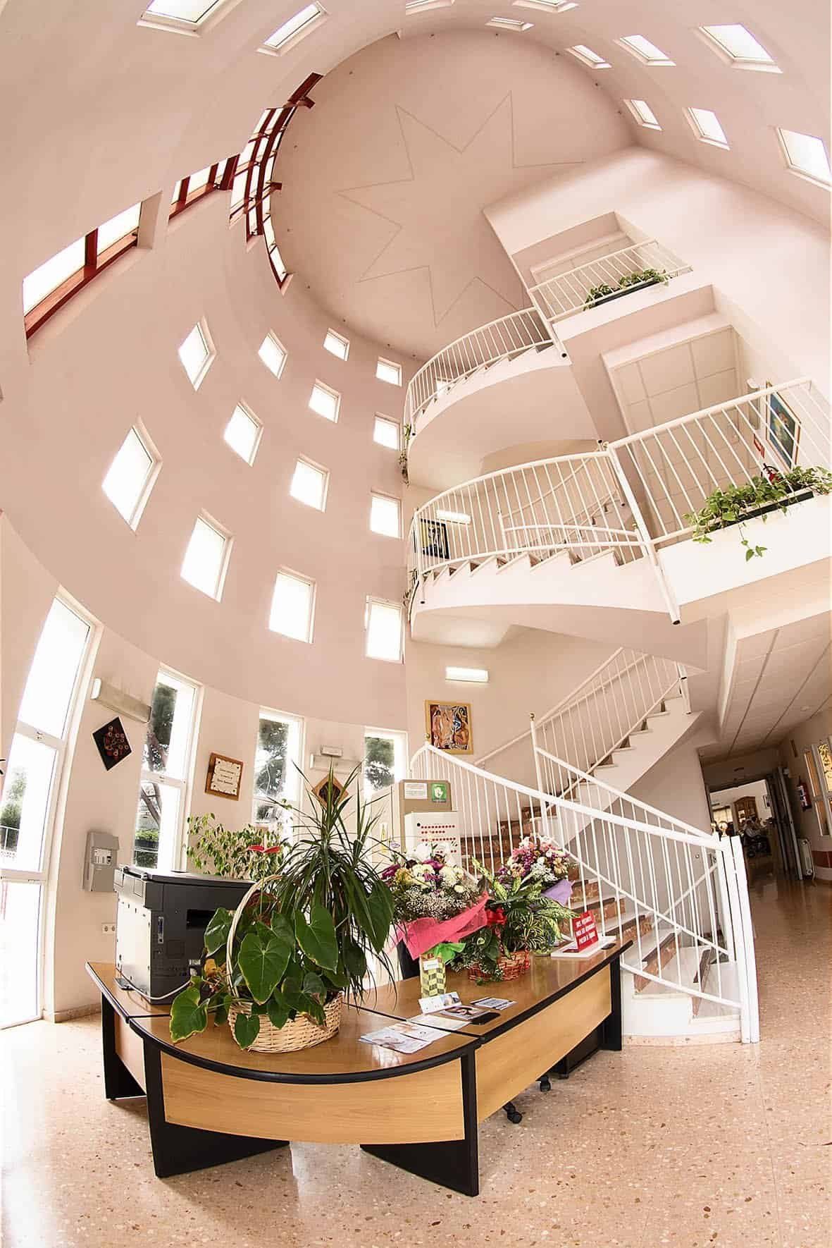 La imagen muestra un espacio interior moderno, luminoso y aireado. La arquitectura es singular, con un diseño circular de varios niveles. Una escalera de caracol prominente con barandillas blancas se curva hacia arriba por el centro del espacio. El techo es muy alto y tiene un patrón de estrella en una cúpula circular. Las paredes están llenas de pequeñas ventanas rectangulares que dejan entrar mucha luz natural. En la planta baja, hay un mostrador de recepción de madera con plantas y arreglos florales. El suelo es de baldosas de color claro.
