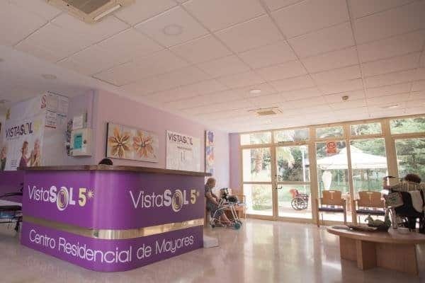 Muestra la recepción de un centro residencial para mayores llamado "VistaSOL 5". Se ve un mostrador curvo de color morado con el logo, una persona mayor sentada con un andador y una zona de espera junto a una gran puerta de cristal que da al exterior.