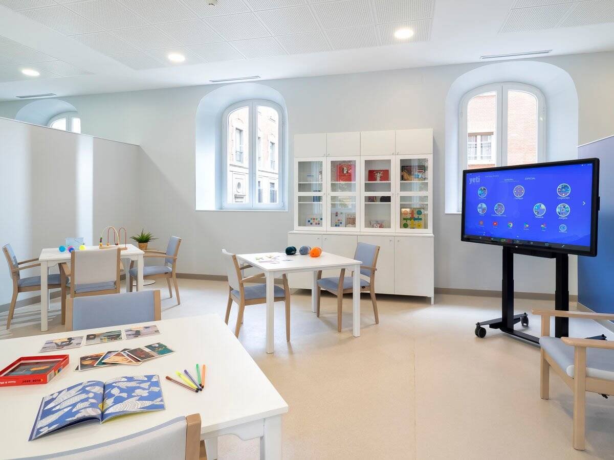 Sala de actividades luminosa de la residencia con mesas, sillas, una televisión de pantalla táctil y grandes ventanas con arcos.