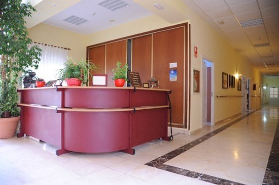 Recepción de la Residencia Mar de Castilla, con un mostrador curvo de color granate, plantas decorativas y un pasillo luminoso al fondo.