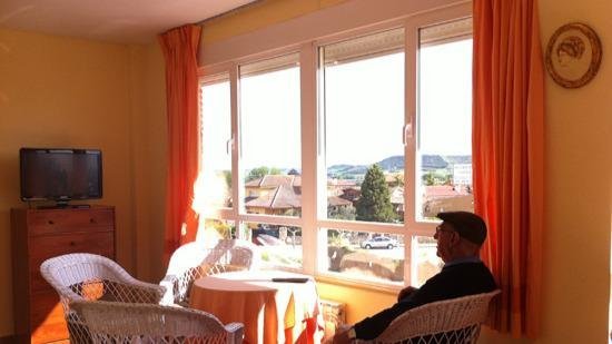 Un residente sentado en un sillón de mimbre en una sala luminosa con paredes amarillas, mirando por una gran ventana con vistas a la ciudad.