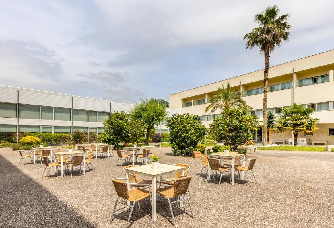 Fachada de la residencia DomusVi Barreiro con una gran terraza exterior, mesas y sillas, rodeada de jardines y palmeras.
