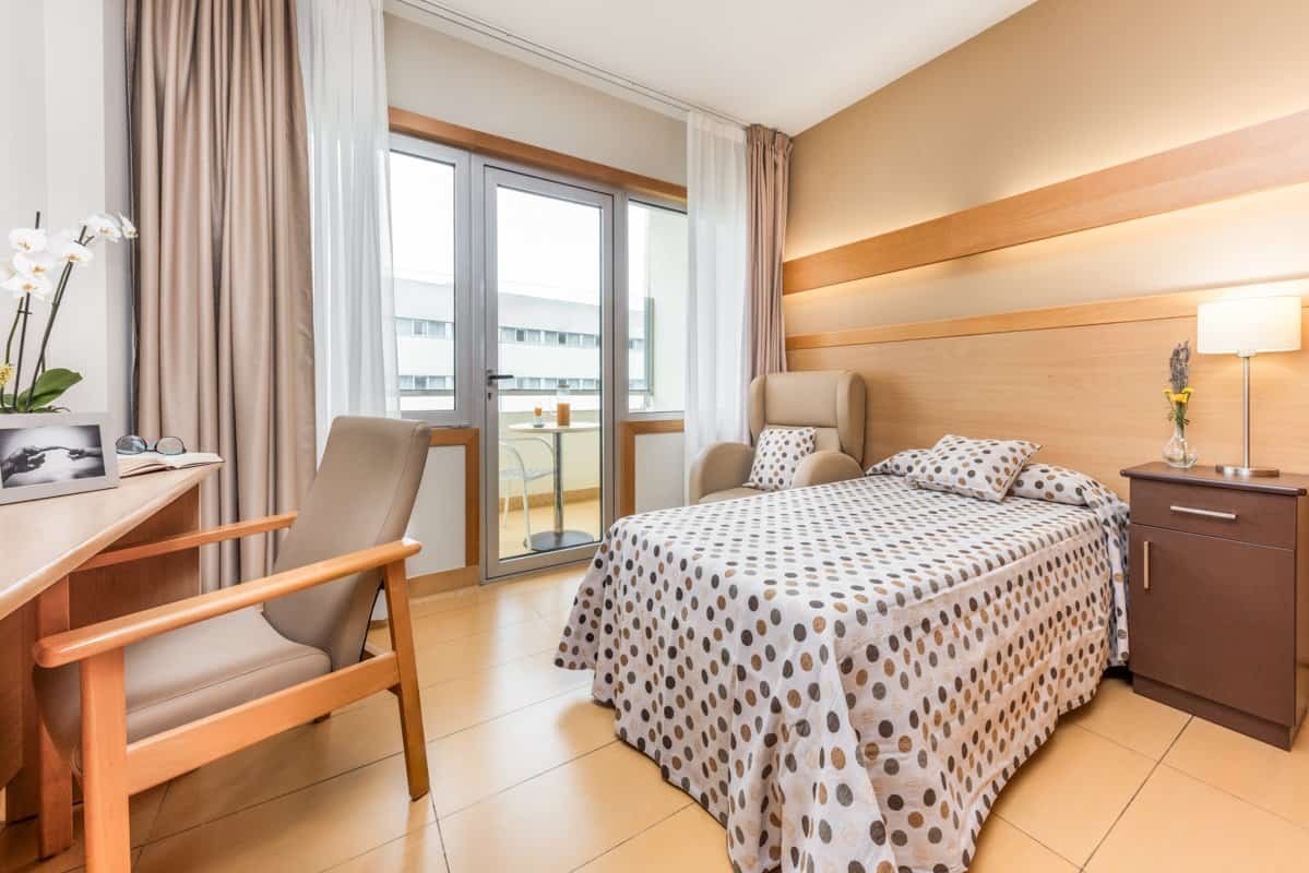 Habitación individual con una cama con ropa de cama de puntos marrones, un escritorio y una puerta de cristal que da acceso a una terraza.