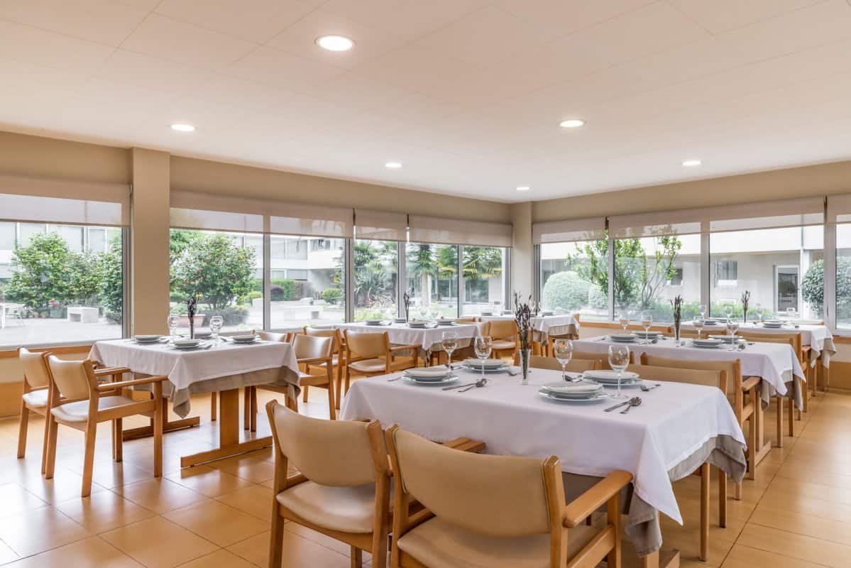 Comedor de la residencia con mesas dispuestas para comer y grandes ventanales con vistas al jardín.