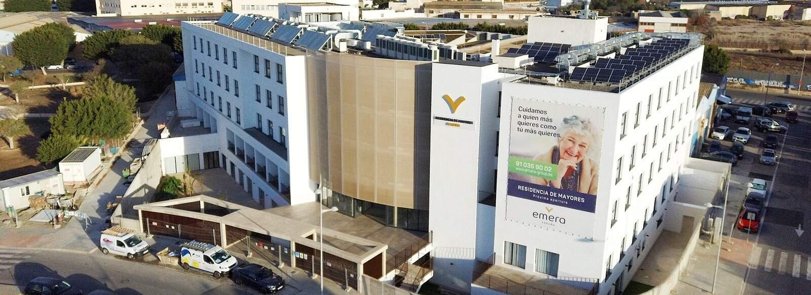 Vista aérea de un complejo de edificios blancos y beige, presumiblemente una residencia de mayores llamada "emera". El tejado de varios de los edificios cuenta con paneles solares. Un gran cartel vertical en uno de los edificios muestra el logo de "emera" y la fotografía de una mujer mayor sonriendo, junto con texto en español y un número de teléfono. Alrededor del complejo se ven zonas ajardinadas, carreteras con coches aparcados y algo de vegetación. El día es soleado.