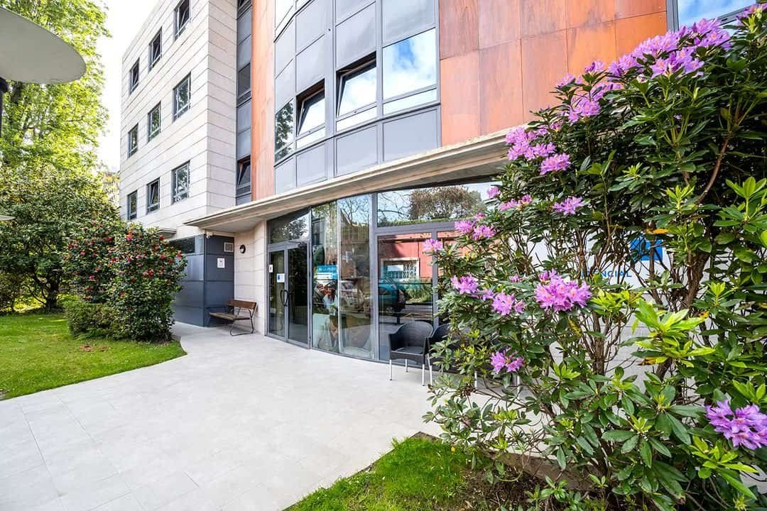 Entrada de la Residencia Sanitas Vigo, un edificio moderno con fachada de cristal y rodeado de un jardín con arbustos y flores rosas.