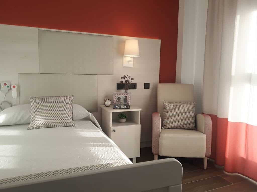 Habitación de residencia confortable y luminosa con cama individual vestida en blanco y cojín estampado, mesita de noche con lámpara, decoración personal y un cómodo sillón a juego. Los tonos cálidos beige y naranja crean un ambiente agradable.