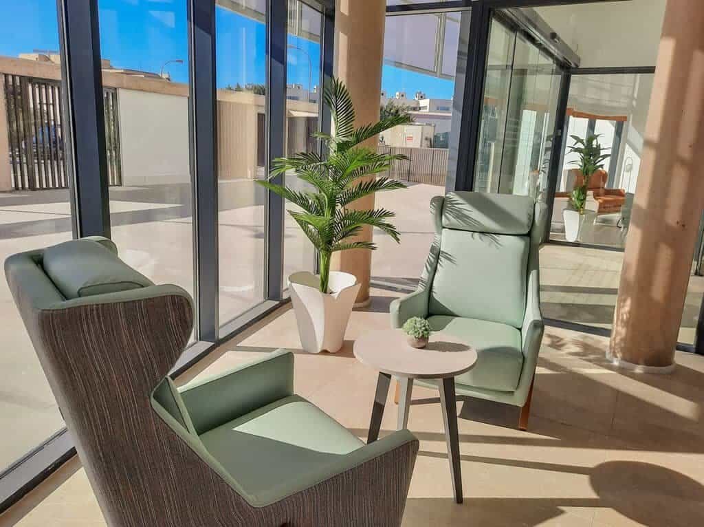 Se presenta una zona de descanso moderna y luminosa. Tres sillones de diferentes estilos (uno gris oscuro y dos verde pálido) están dispuestos alrededor de una pequeña mesa redonda de color claro con una pequeña planta. Un gran ventanal con pilares de color claro permite la entrada de abundante luz natural y ofrece vistas de un exterior soleado con vegetación y otros edificios. Una palmera de interior en una maceta blanca añade un toque de verdor al espacio. El suelo es de baldosas de color beige claro.