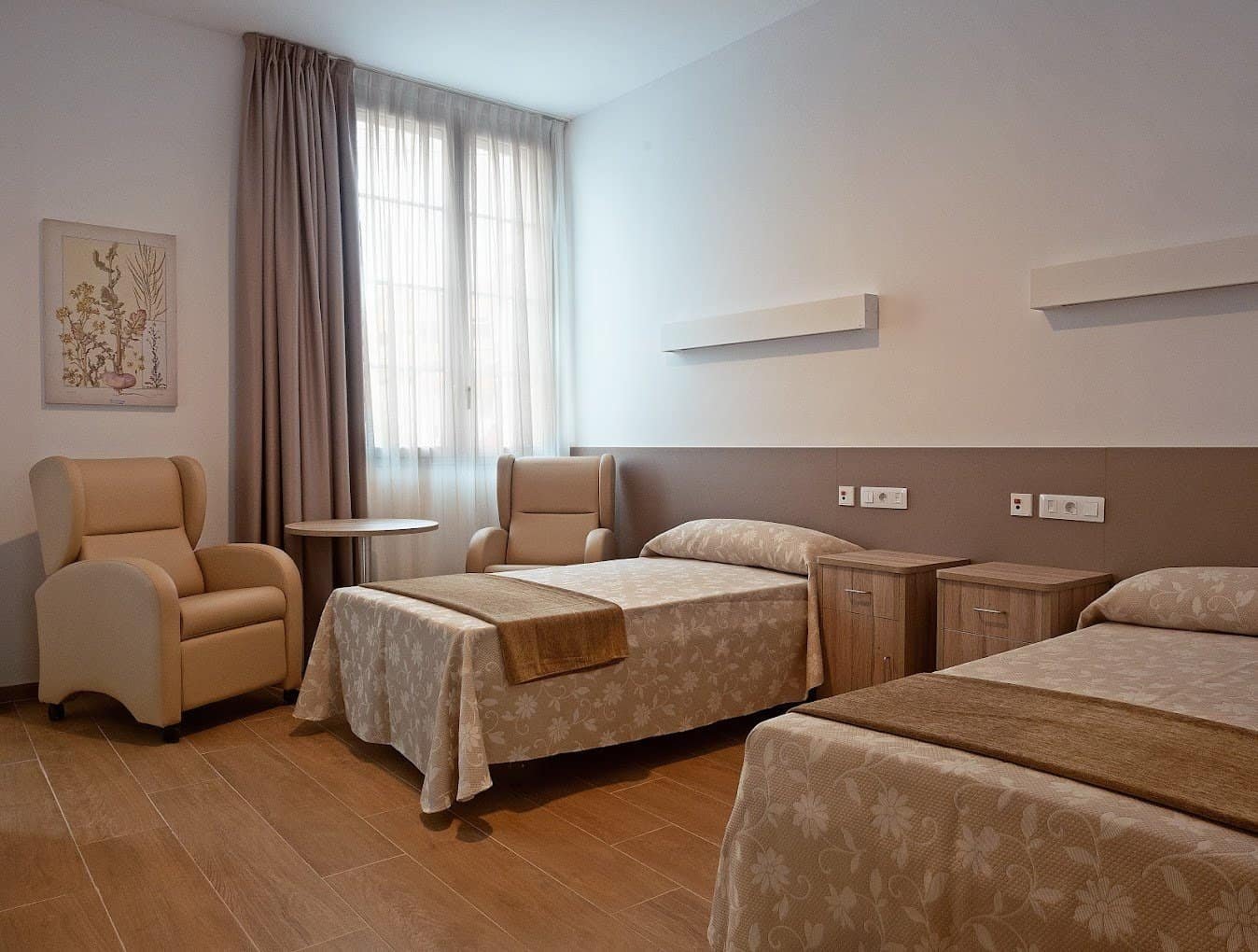 Habitación doble con dos camas, dos sillones color beige, un suelo de madera y una ventana con cortinas.