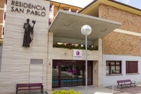 Entrada de la Residencia San Pablo de Vitalia Home en Teruel, con una estatua en la fachada y un banco en el exterior.