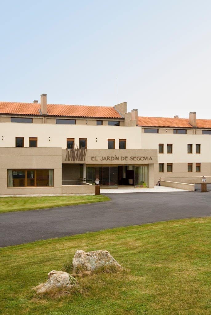 Fachada de la residencia El Jardín de Segovia, un edificio con tejado de tejas rojas, entrada principal moderna y un amplio jardín en la parte delantera.