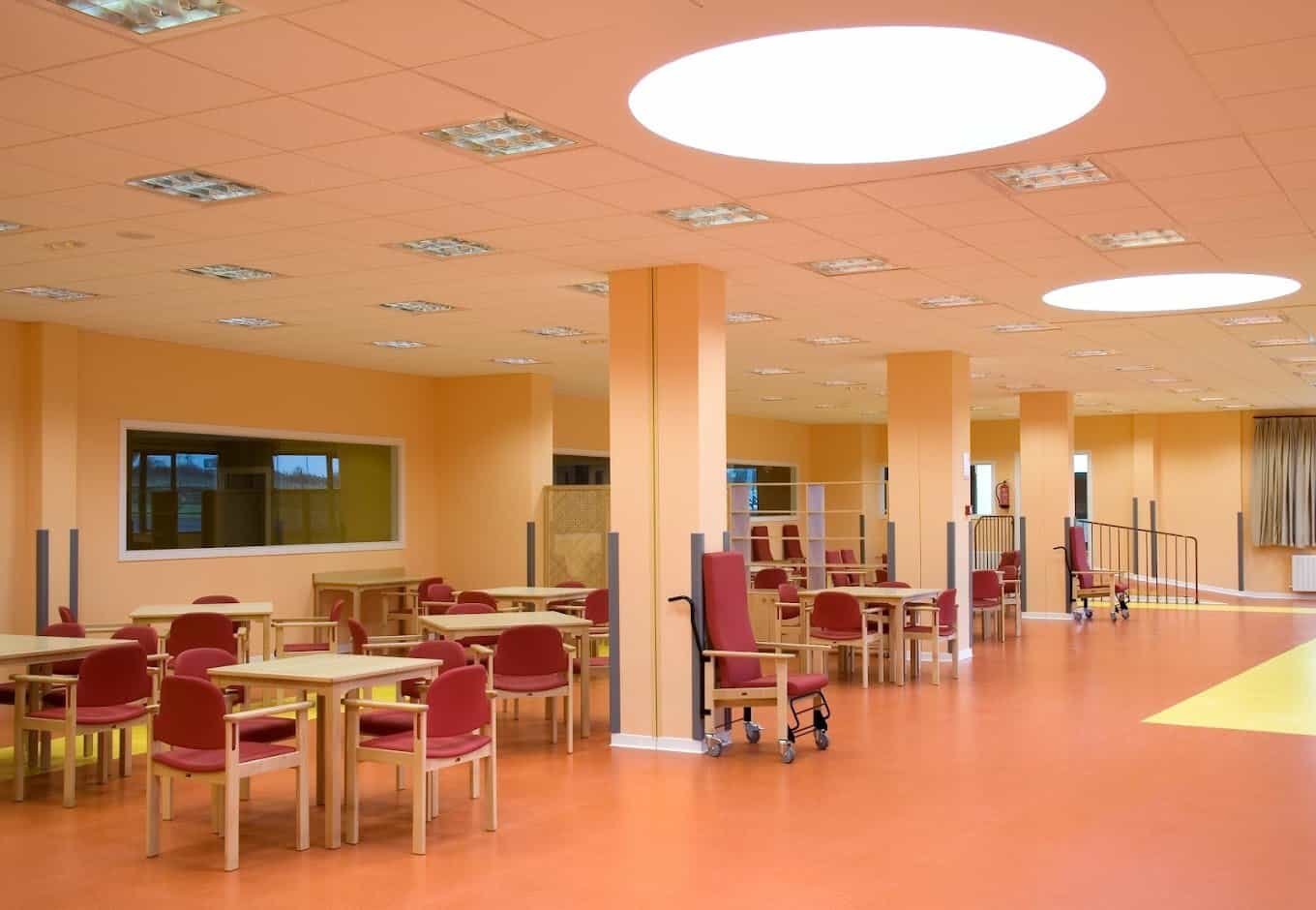 Comedor y sala común de la residencia, con mesas de madera, sillas rojas y un ambiente luminoso y acogedor.