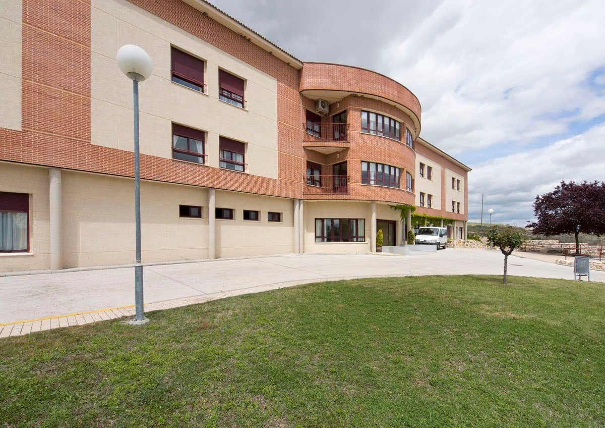 Fachada exterior de la Residencia Virgen del Bustar, un edificio de ladrillo y hormigón con una sección curva y un jardín delantero.