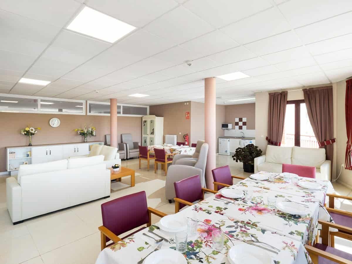 Amplia sala común de la residencia con un gran sofá blanco, sillones, mesas de comedor y un ambiente hogareño.
