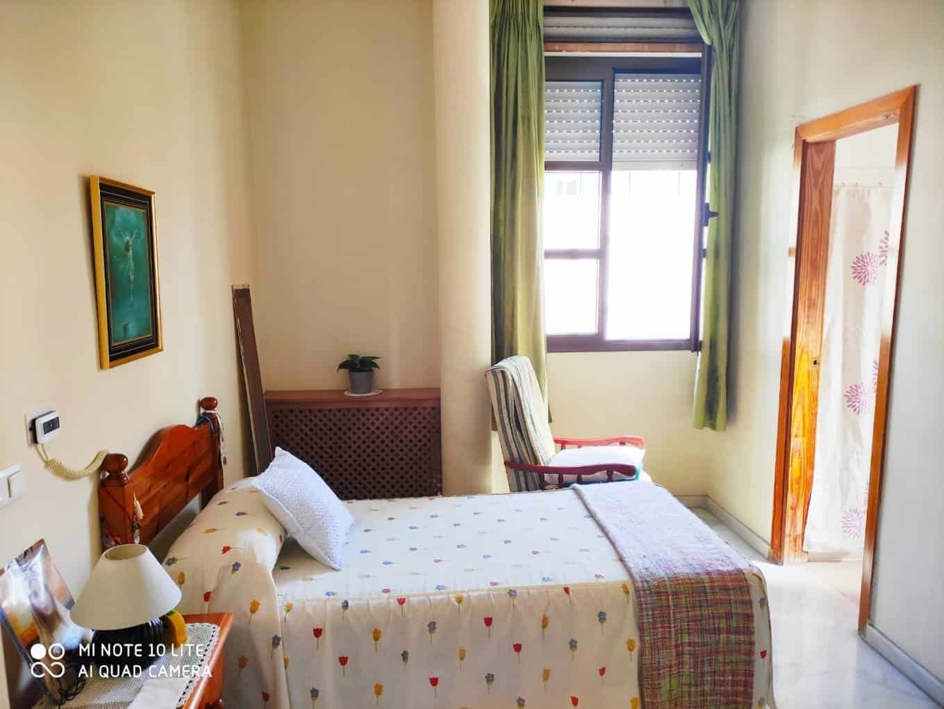 Habitación individual con paredes claras, cama con colcha estampada, un sillón de rayas y una ventana con cortinas verdes.