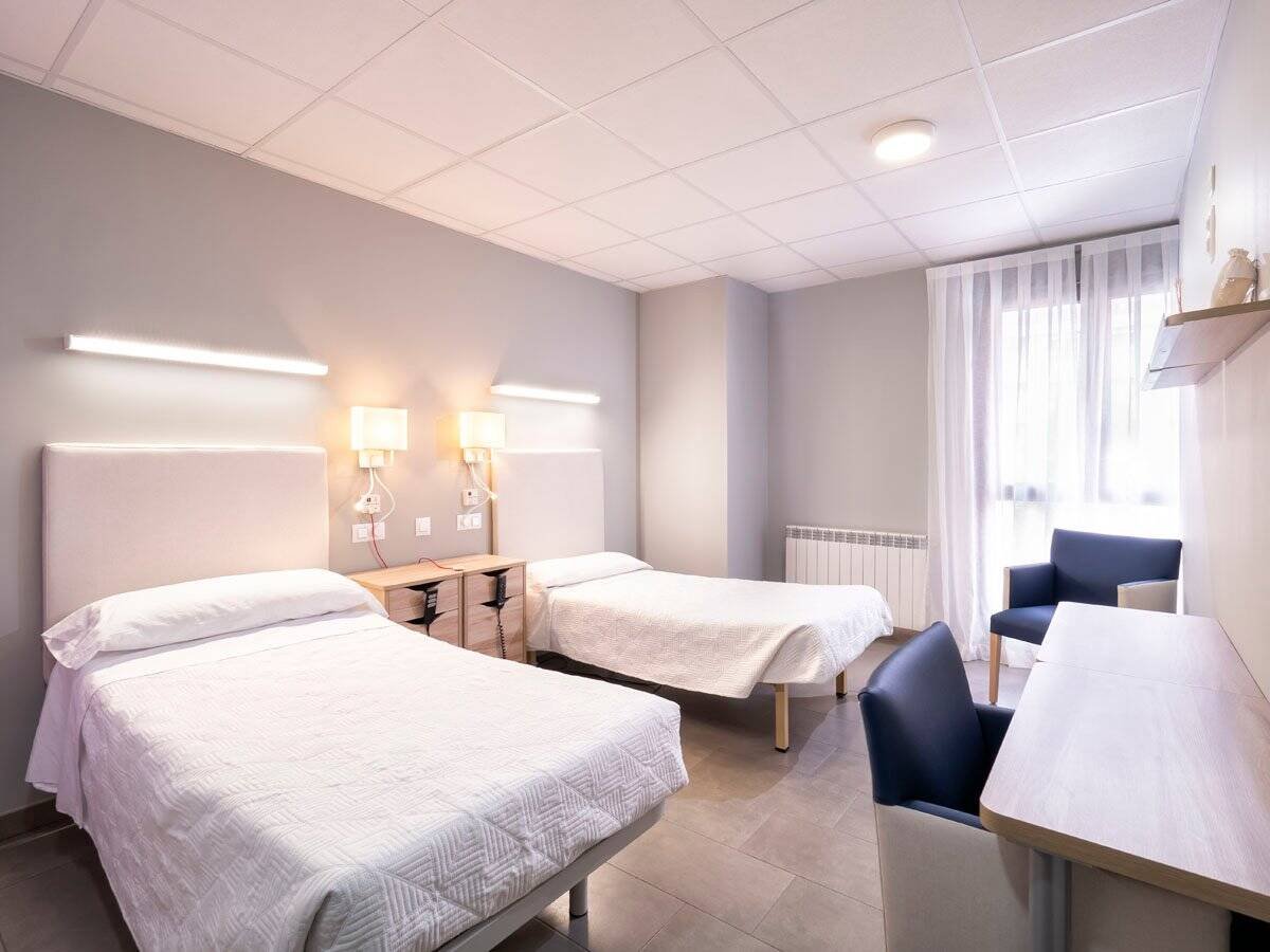 Habitación doble moderna en la Residencia CleceVitam El Parque, con dos camas, escritorio y un gran ventanal.
