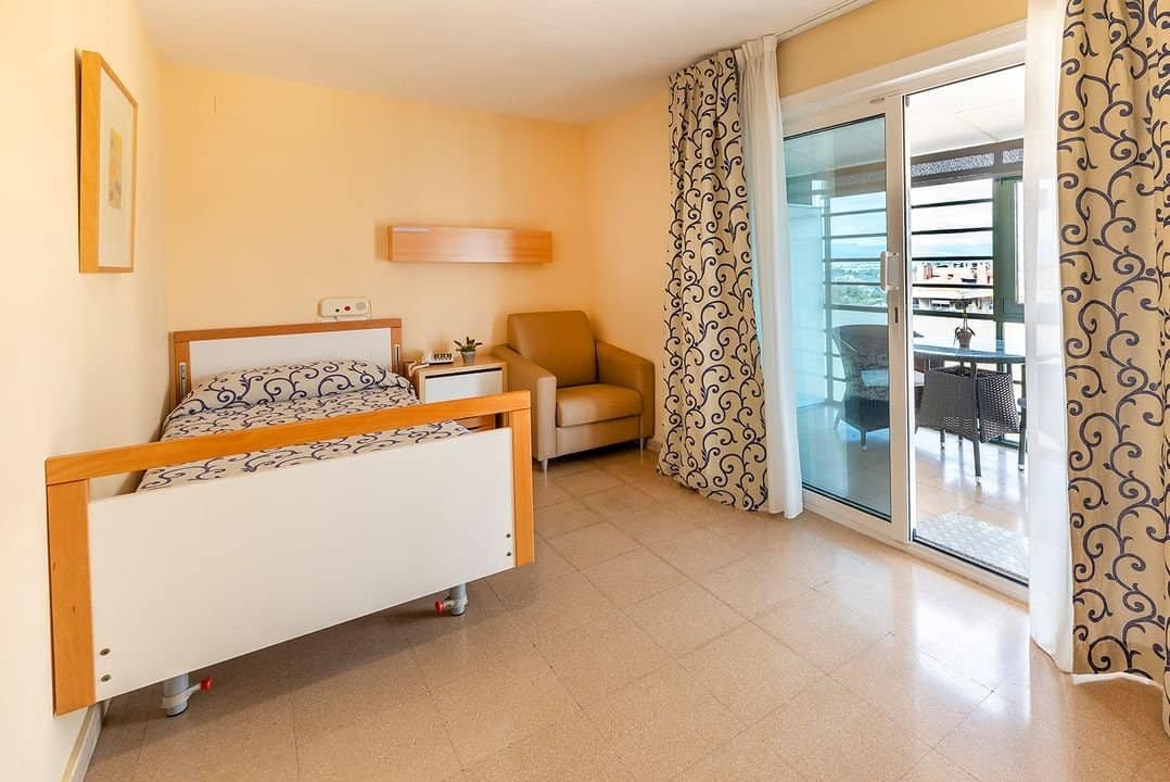 Habitación individual luminosa con cama, sillón y una puerta de cristal que da a una terraza.