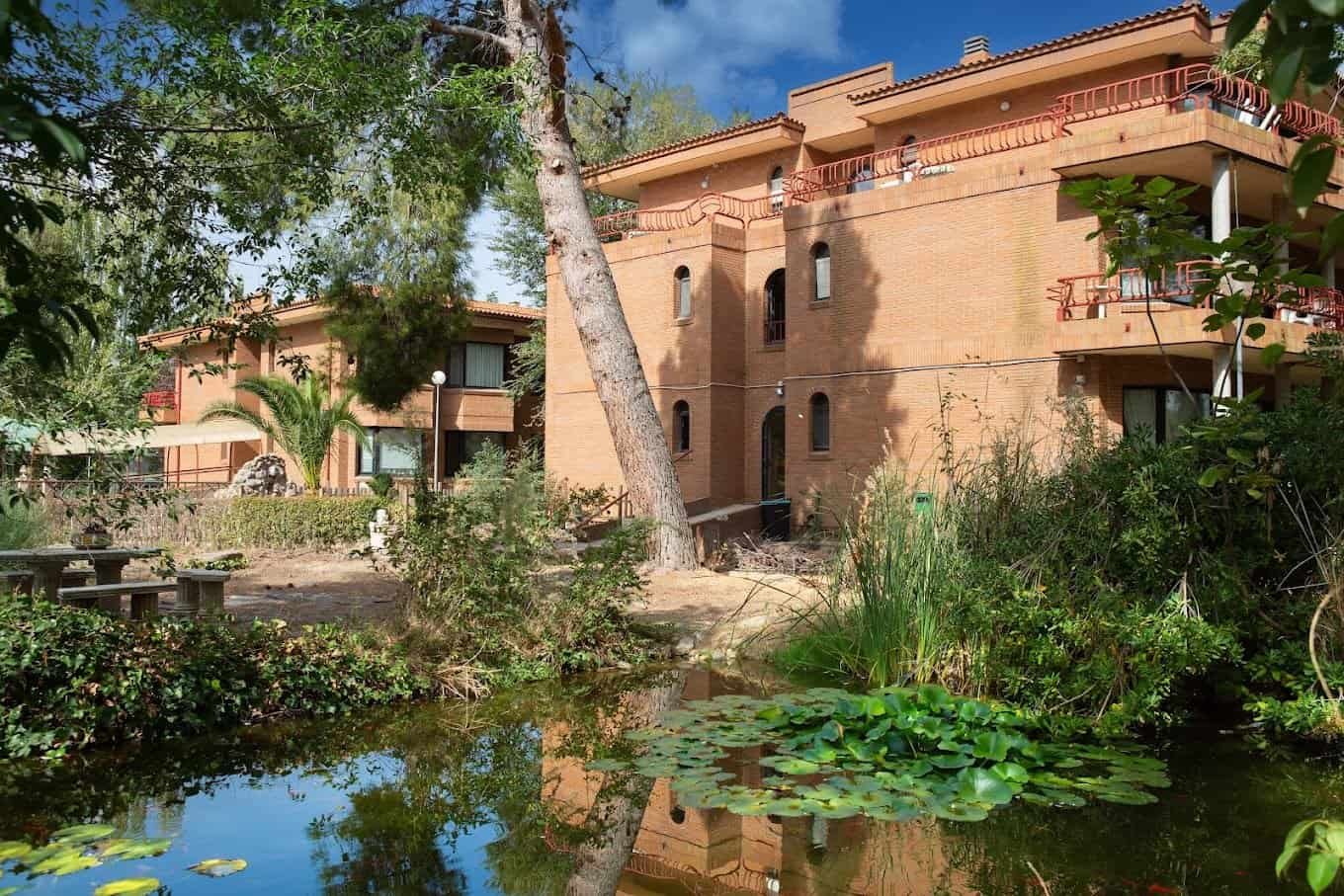 Edificio de ladrillo de la residencia DomusVi Virgen de la Blanca, rodeado de un jardín con estanque, nenúfares y un gran árbol.