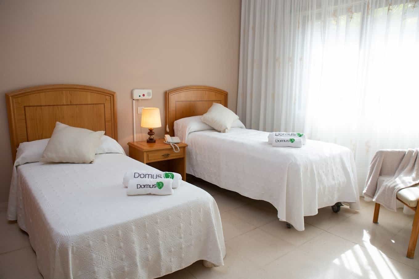 Habitación doble luminosa y limpia en la residencia DomusVi, con dos camas individuales y toallas con el logo de la marca.