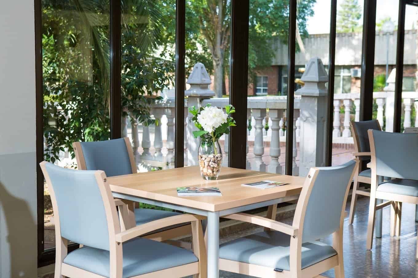 Zona de estar interior en la residencia DomusVi con una mesa, sillas y una ventana con vistas a un jardín con balaustrada.