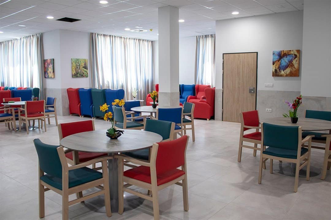 Espaciosa sala común de Vitalia Home con mesas redondas, sillas de colores, sillones y cuadros en las paredes.