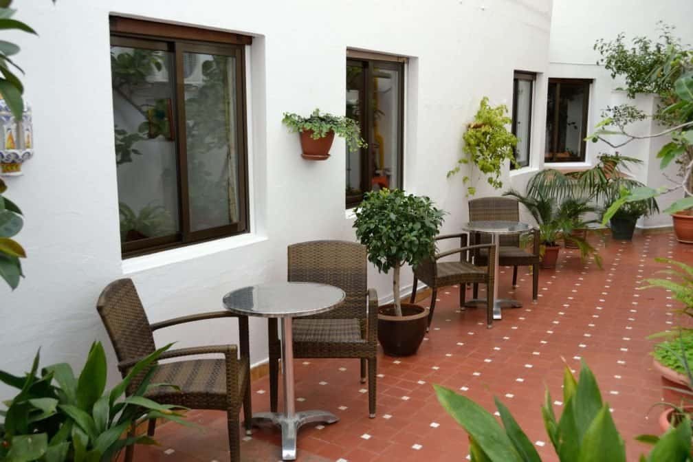 Patio interior de la residencia con suelo de azulejos, mesas redondas, sillas de mimbre y plantas en macetas.