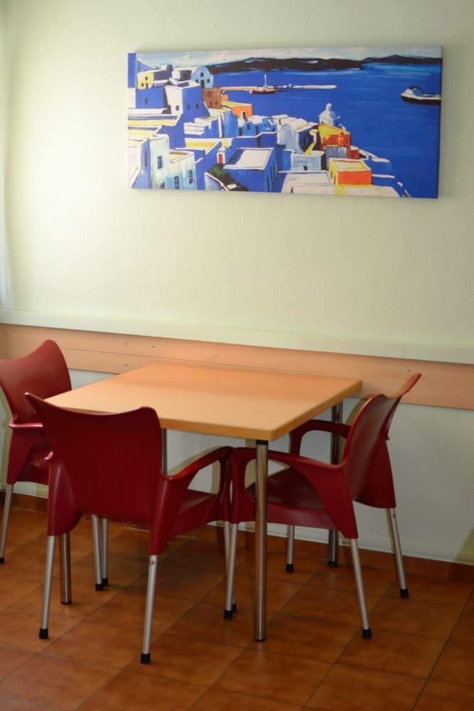 Área de comedor en la residencia con una mesa cuadrada de madera, cuatro sillas rojas y un cuadro grande de una ciudad costera.
