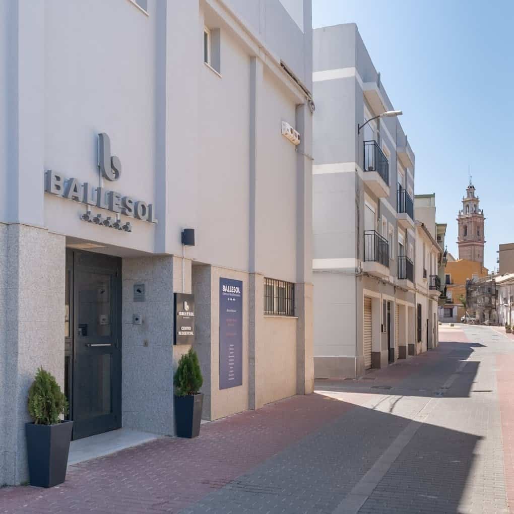 Fachada exterior de la Residencia Ballesol Burjassot, con el logo de la marca en la entrada principal y una torre de iglesia visible al fondo.