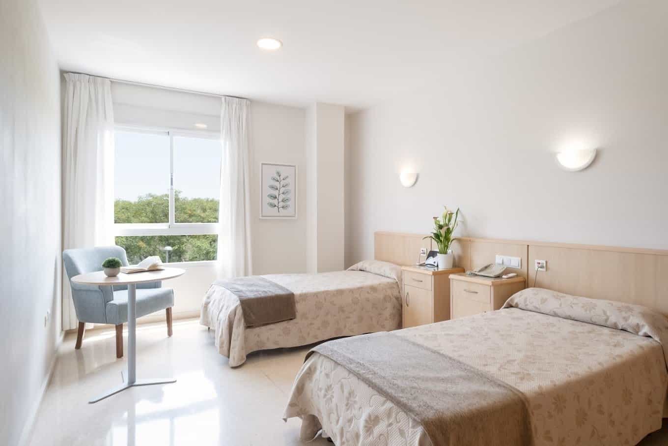 Habitación doble luminosa y limpia en la Residencia Ballesol, con dos camas individuales, una silla y una mesa pequeña junto a la ventana.