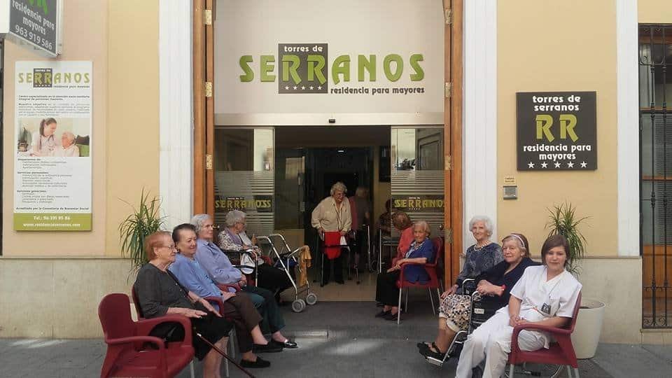 Fachada de la Residencia Torres de Serranos en Valencia con personas mayores y una cuidadora sentadas en sillas rojas.