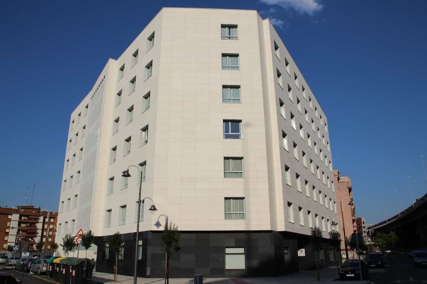 Edificio moderno de seis pisos con fachada blanca y base oscura, ubicado en una esquina de la calle, con grandes ventanales y árboles pequeños en la acera.