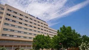 Vista exterior del gran edificio de varias plantas de la residencia Albertia Valdespartera, con árboles verdes en primer plano bajo un cielo azul.