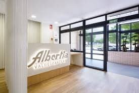 La moderna y luminosa recepción de la residencia Albertia Valdespartera, con un mostrador blanco que muestra el logo iluminado y un suelo de madera clara.