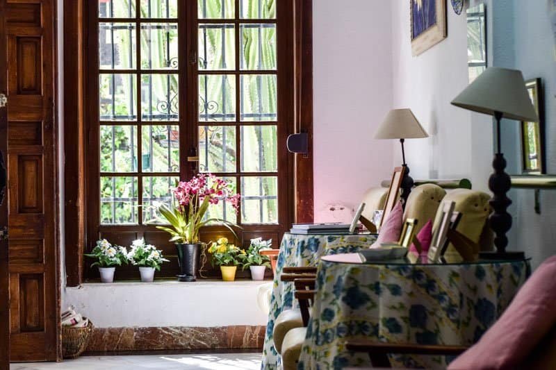 Rincón de una sala de estar en Residencia Aurora que muestra una gran ventana de madera con plantas en el alféizar y mesas cubiertas con manteles florales en primer plano.