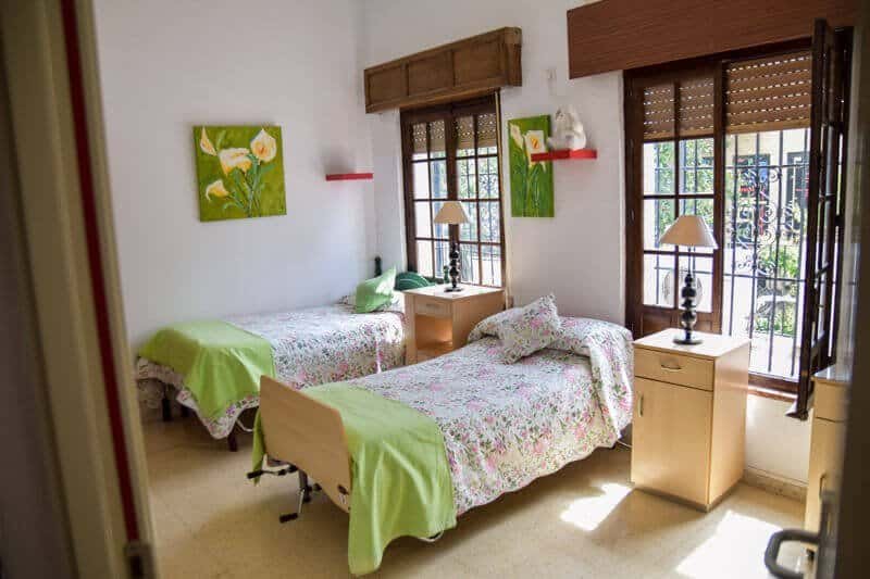 Habitación doble, luminosa y hogareña en Residencia Aurora. Cuenta con dos camas con edredones florales, mobiliario de madera y grandes ventanas con contraventanas.