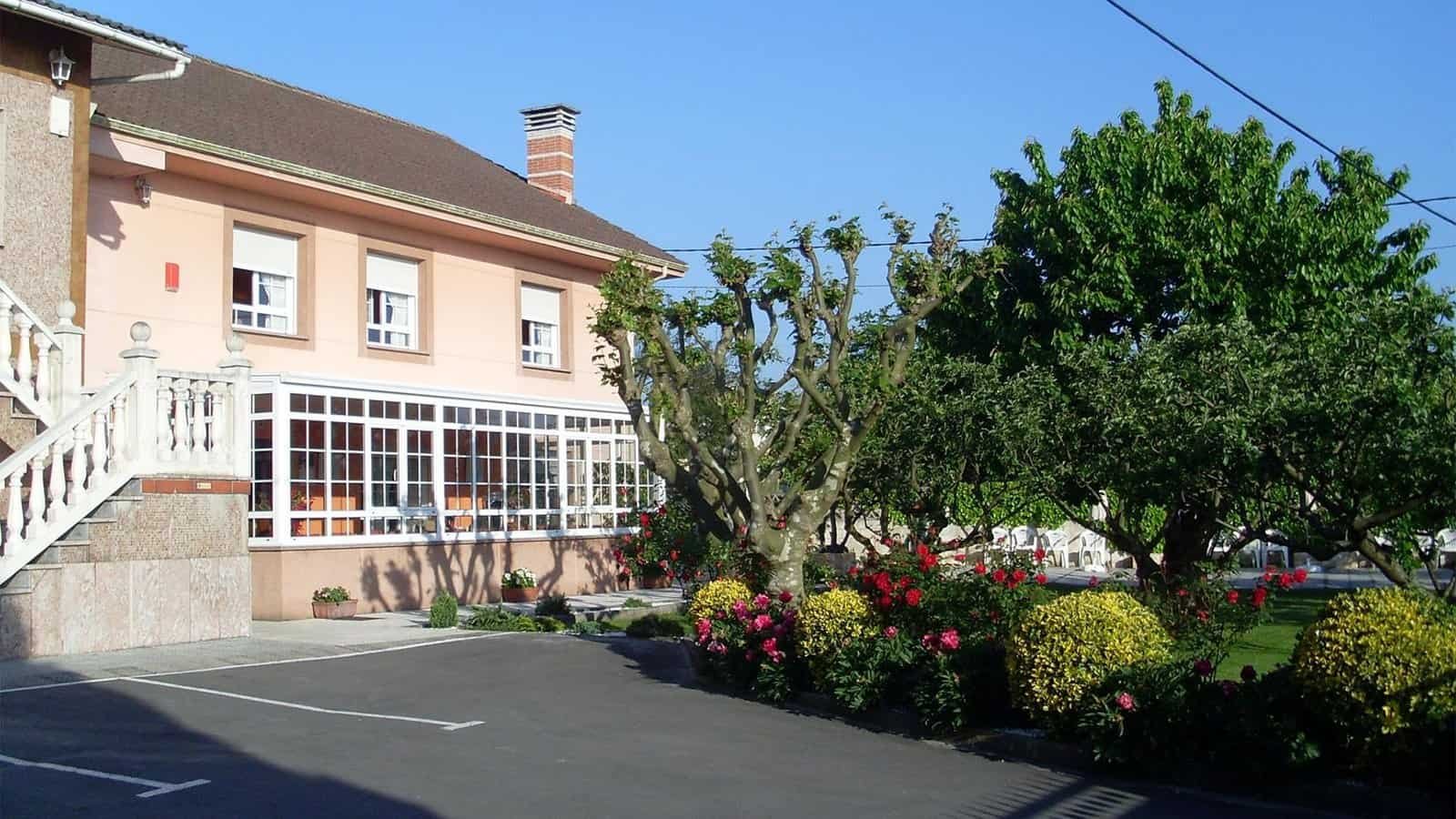 Exterior de una residencia con fachada color melocotón, un gran ventanal que da a una sala acristalada, y un jardín bien cuidado con árboles y flores.