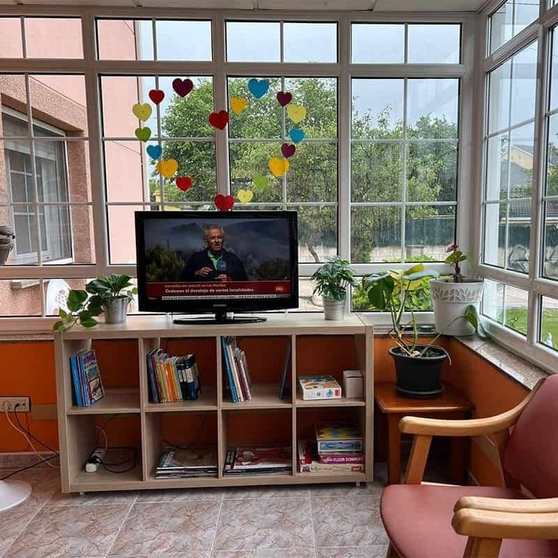 Interior de una sala acristalada con un televisor sobre un estante lleno de libros y juegos. La sala está decorada con un corazón de papel de colores.