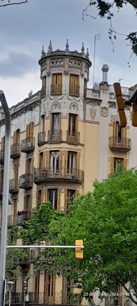 Un primer plano de la fachada ornamentada de un edificio de estilo modernista con una torre distintiva, balcones de hierro forjado y detalles decorativos en tonos claros. Se ven árboles verdes en la parte inferior de la imagen.