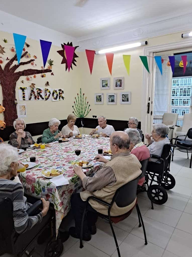 Un grupo de personas mayores están sentadas alrededor de una mesa cubierta con un mantel floral, disfrutando de una comida en una sala decorada con motivos otoñales y guirnaldas de colores. Algunas personas están en sillas de ruedas.