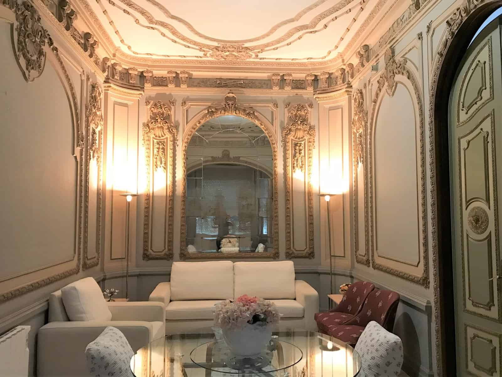 Una sala de estar elegante y ornamentada con paredes decoradas con molduras clásicas, dos sofás de color claro, una mesa de centro de cristal y espejos que amplían visualmente el espacio.