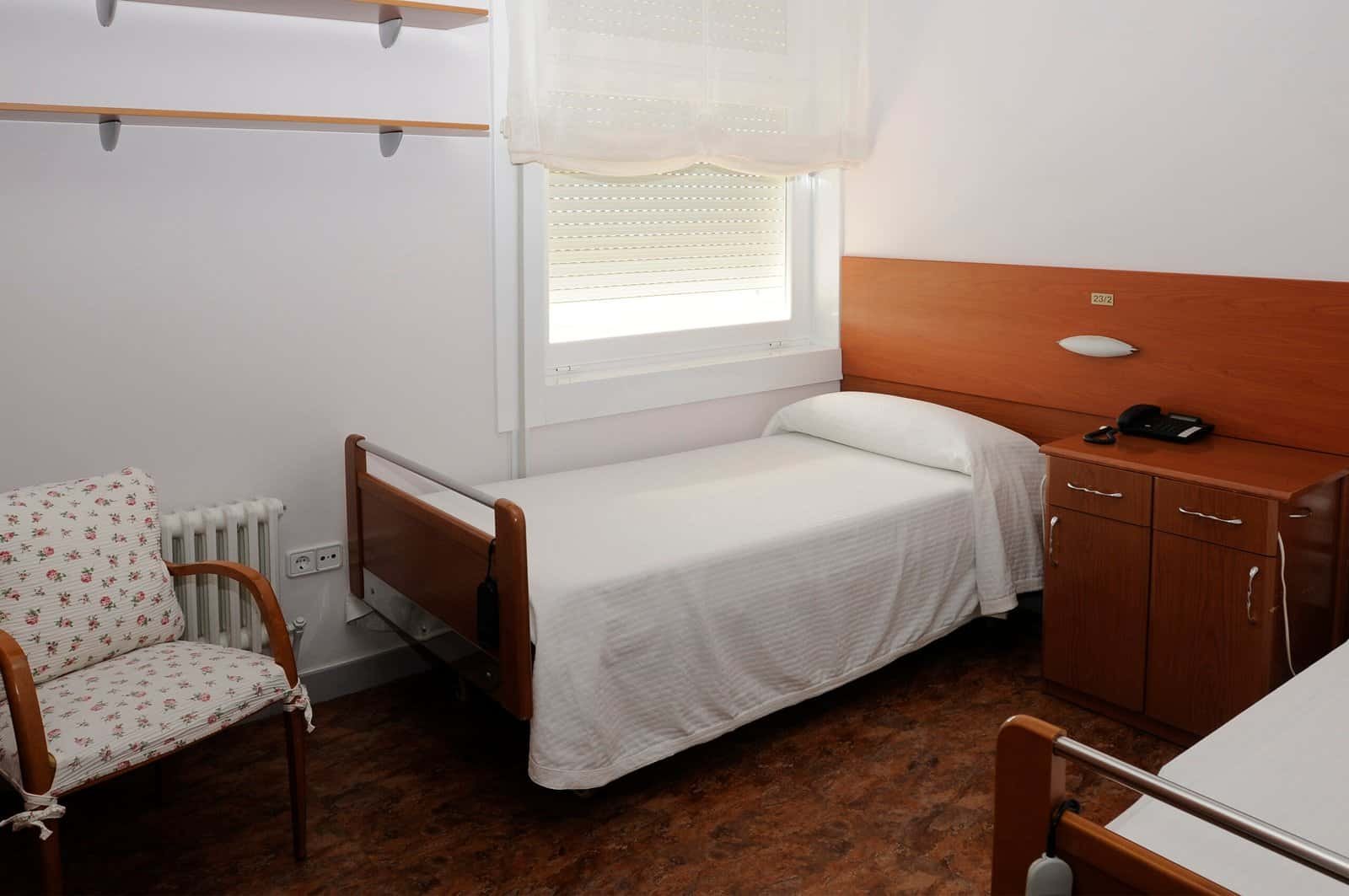 Una habitación sencilla con dos camas individuales con ropa de cama blanca, una mesita de noche de madera con un teléfono, una silla con estampado floral y una ventana con una persiana blanca.