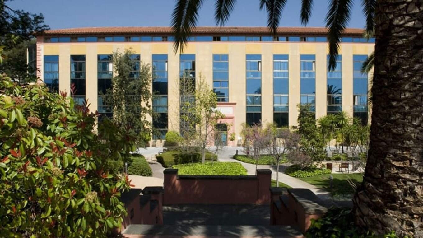La fachada de un edificio de varios pisos en tonos amarillos, con numerosas ventanas azules y un tejado rojizo. Delante del edificio hay vegetación exuberante, incluyendo árboles y palmeras.