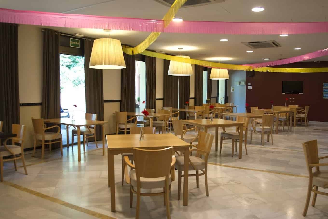 Un comedor amplio y luminoso con mesas de madera clara y sillas con tapicería beige. El techo está decorado con guirnaldas festivas de color rosa y amarillo.