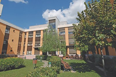 Vista exterior de un edificio moderno de varios pisos en tonos naranja y beige, con amplias ventanas y rodeado de zonas ajardinadas con césped, árboles y flores. El cielo está parcialmente nublado.
