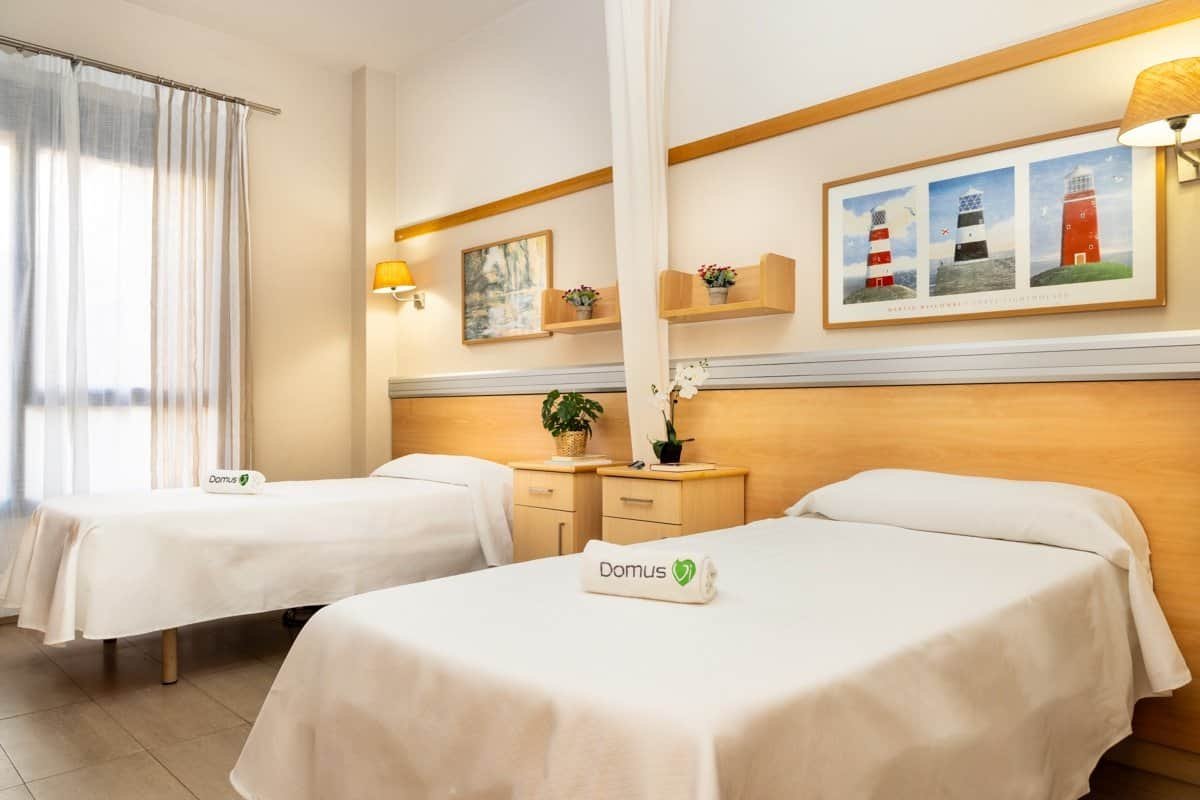 Una habitación doble luminosa y moderna en una residencia, con dos camas individuales con ropa de cama blanca, mesitas de noche de madera clara y decoración sencilla incluyendo lámparas y cuadros.