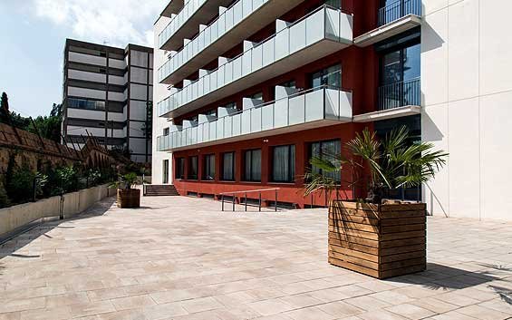 Fachada de un edificio moderno con balcones de cristal y una terraza con macetas de plantas, en un entorno urbano.