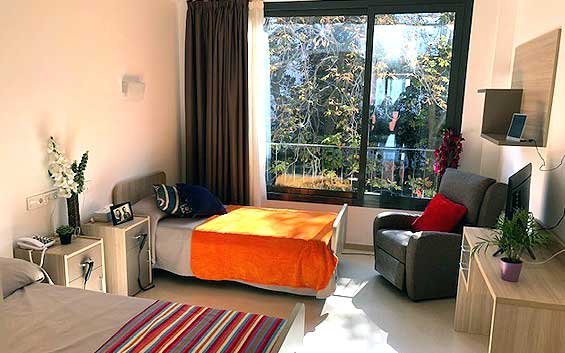 Habitación de una residencia con dos camas individuales, un sillón gris y un gran ventanal con vistas a los árboles.